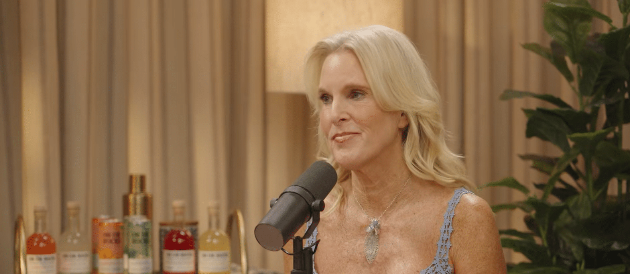 A woman with blonde hair speaks into a microphone in a studio setting, highlighting the art of video production. Bottles and a plant are visible in the background, and warm lighting creates a cozy atmosphere.